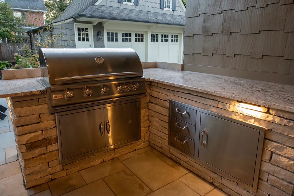 Built-in grill wall with stainless grill, storage drawers, natural stone, countertop, and under-counter lighting